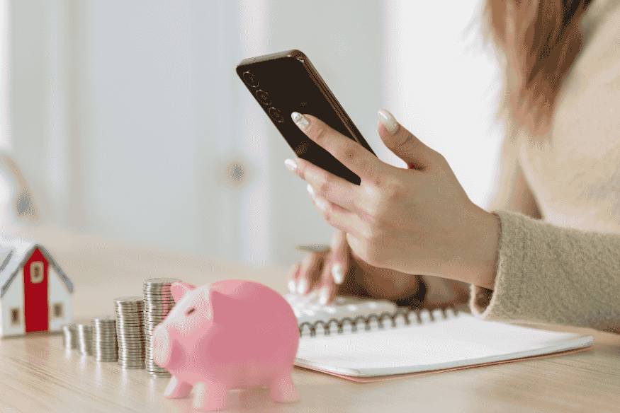 Mulher utilizando aplicativo de finanças no celular ao lado de cofrinho rosa e moedas empilhadas, representando organização financeira para aumentar o score de crédito.