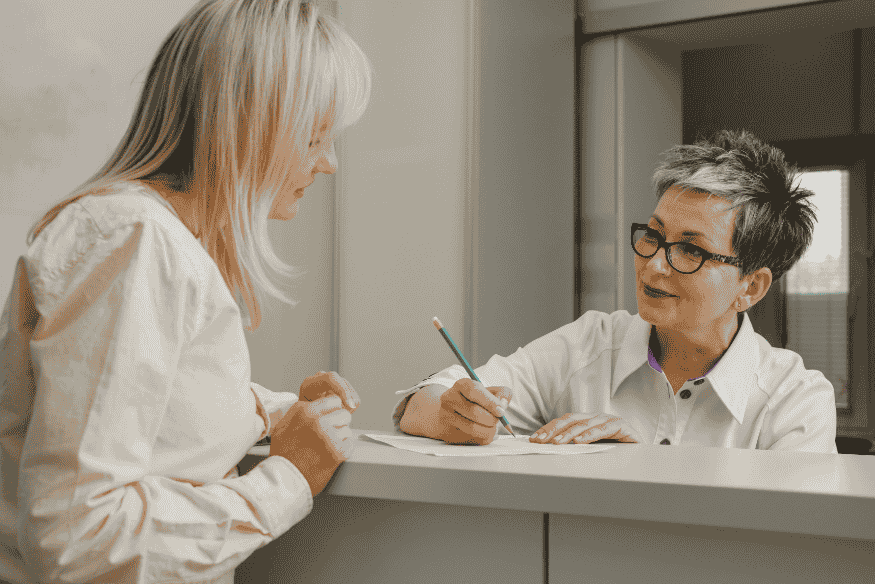 Duas mulheres conversando em um balcão, com uma delas preenchendo documentos, simbolizando o processo de ajuste e atualização de informações no Cadastro Positivo.