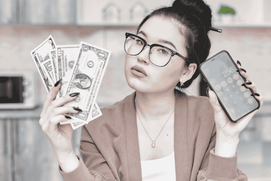 Mulher segurando notas de dólar e celular com calculadora, simbolizando a análise de variações nos valores das parcelas em financiamentos com taxas variáveis.
