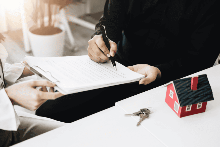 Pessoa assinando contrato de financiamento com chave e miniatura de casa na mesa, representando dívidas com garantia de bens como imóveis e veículos.