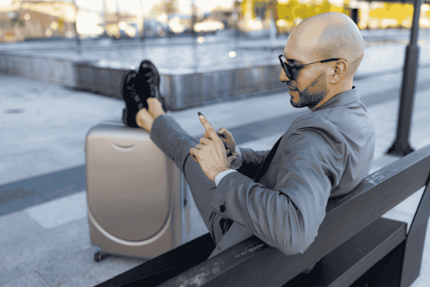 Homem de terno relaxando em banco público com mala de viagem, usando o celular para acompanhar sua saúde financeira e a evolução do score de crédito após quitar dívidas.