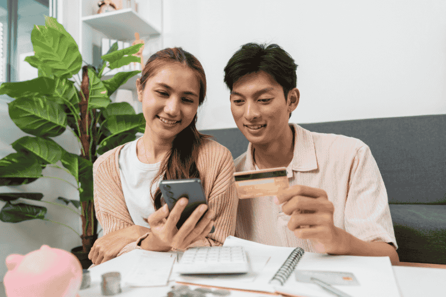 Casal sorrindo enquanto olha para o celular e um cartão de crédito, simbolizando a confiança e controle financeiro, resultado de um bom score de crédito.