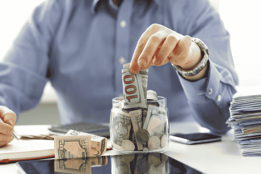 Homem colocando notas de dólar em um pote de vidro, representando o uso inteligente da renda extra, como 13º salário e férias, para quitar dívidas e aliviar o orçamento.