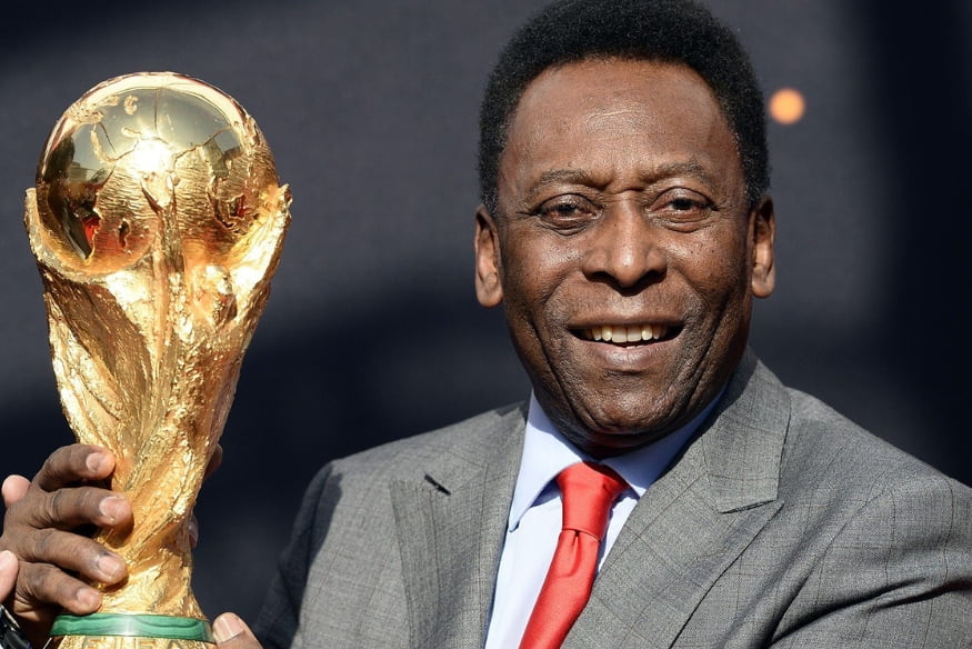 Pelé sorrindo e segurando a taça da Copa do Mundo com terno cinza e gravata vermelha.