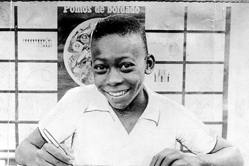 Foto em preto e branco de Pelé criança sorrindo e escrevendo em caderno.