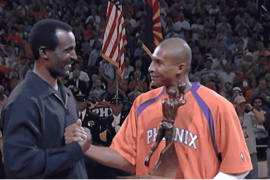 Leandrinho Barbosa segura troféu de prêmio individual da NBA enquanto cumprimenta outro ex-jogador durante cerimônia.