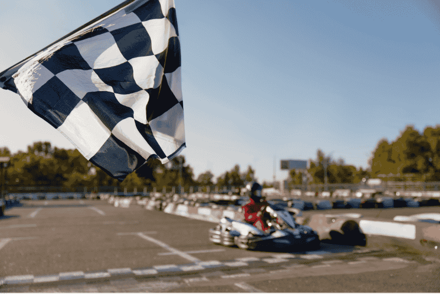 Bandeira quadriculada sendo agitada na linha de chegada de uma pista de kart em ambiente urbano.