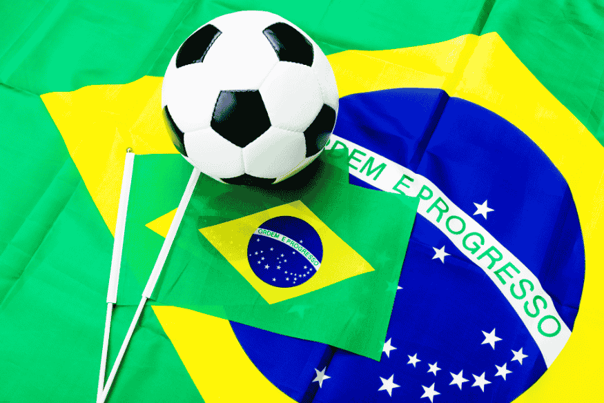 Bola de futebol sobre bandeiras do Brasil em fundo verde e amarelo.
