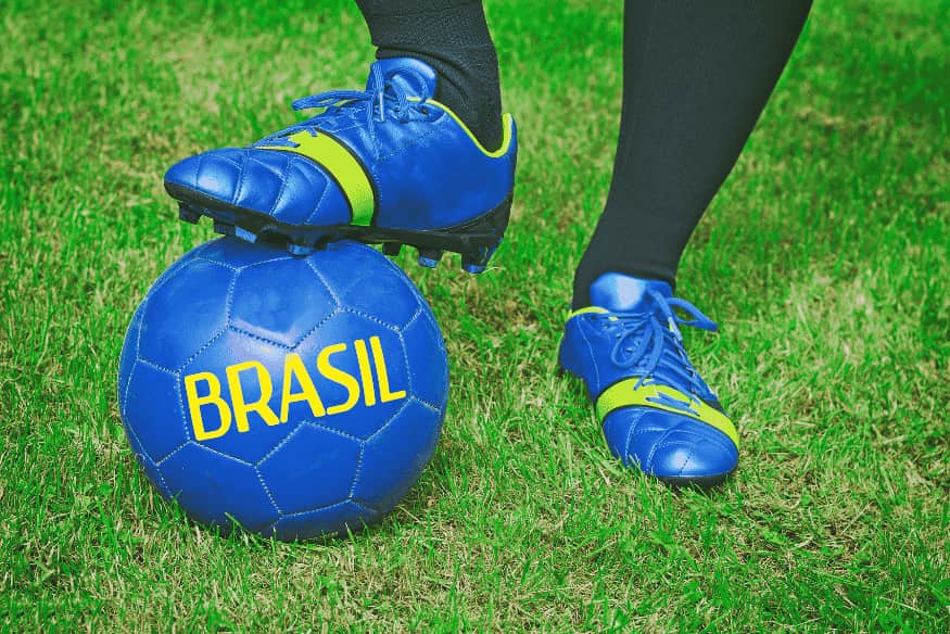 Jogador com chuteira azul apoiada sobre bola azul com a palavra “Brasil”, em campo gramado.