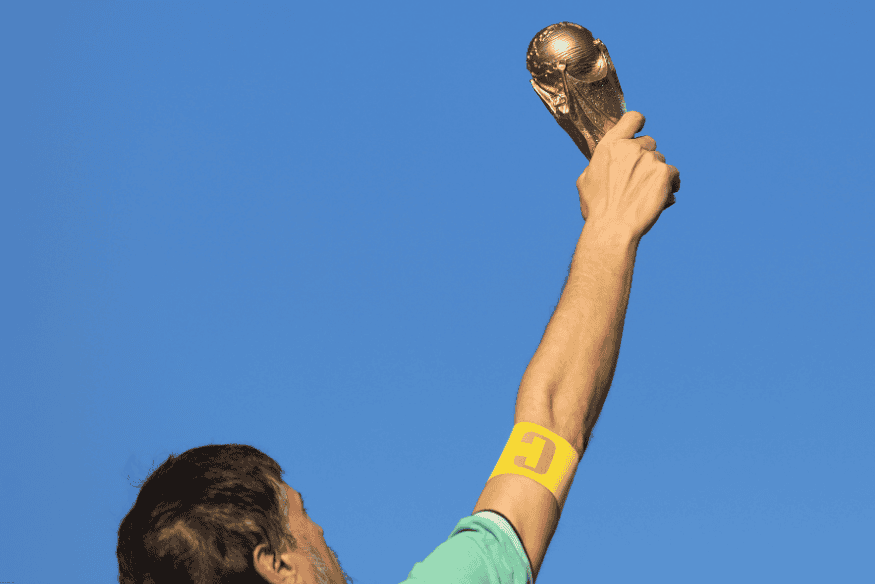 Jogador com faixa de capitão levanta troféu dourado com o céu azul ao fundo.