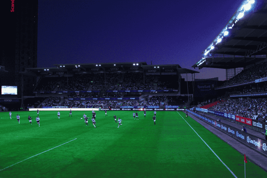 Partida de futebol acontecendo à noite em estádio moderno com iluminação artificial.