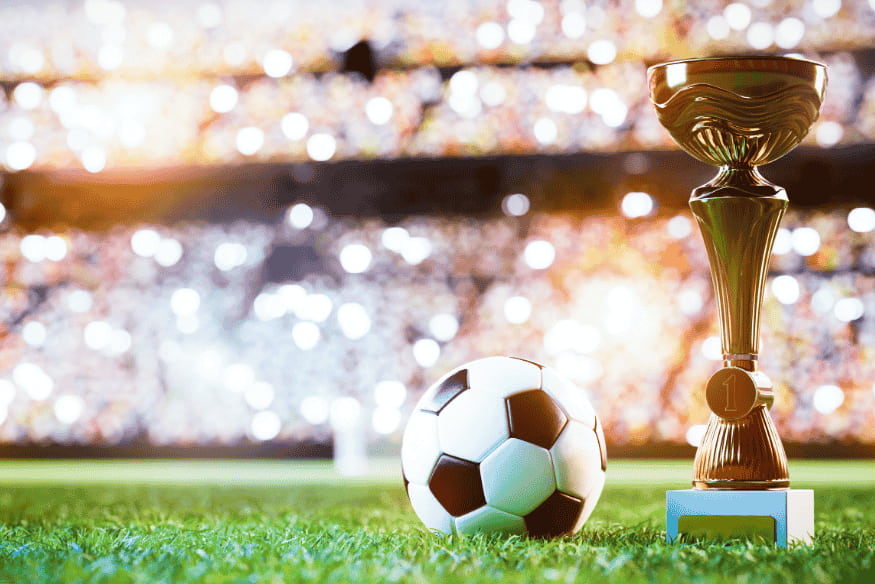 Troféu de futebol dourado ao lado de uma bola em campo, com estádio iluminado ao fundo.