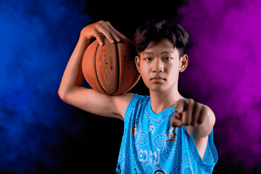 Jovem jogador posa com bola de basquete, apontando para a câmera com expressão determinada.