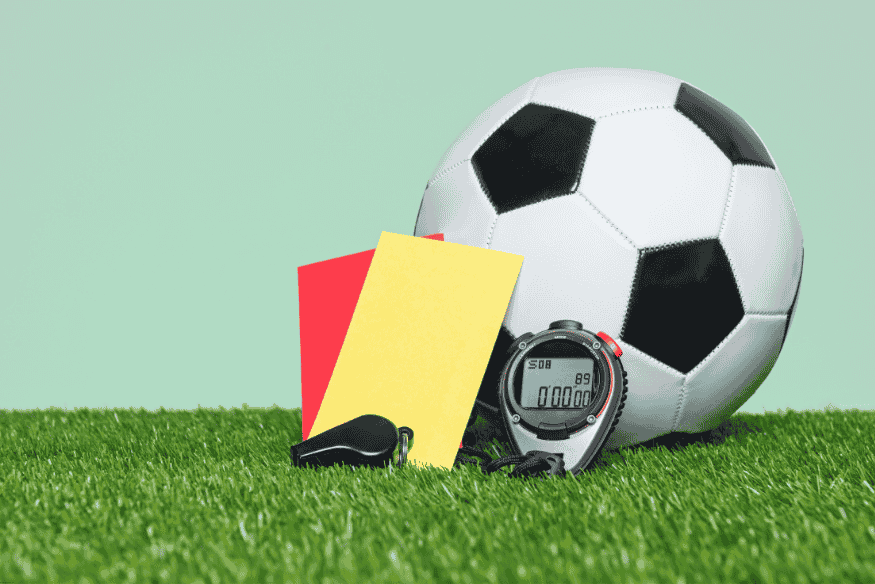 Bola de futebol ao lado de cartões vermelho e amarelo, apito e cronômetro sobre a grama.