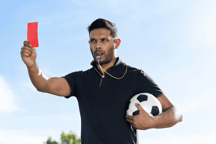 Árbitro de futebol assoprando apito e mostrando cartão vermelho, segurando uma bola.