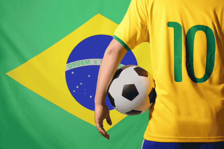Jogador de camisa amarela número 10 segura bola de futebol em frente à bandeira do Brasil.