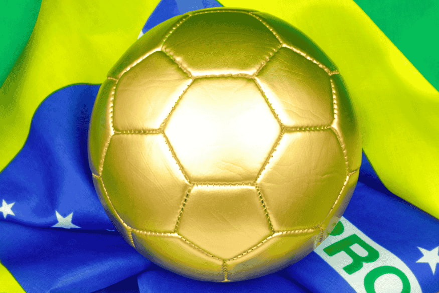 Bola de futebol dourada posicionada sobre a bandeira do Brasil.