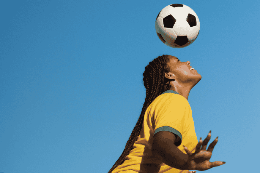 Mulher com camisa amarela controlando bola de futebol com a testa, sob céu azul.