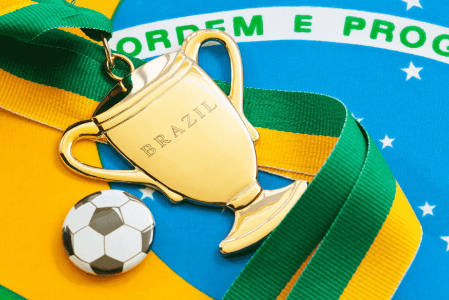 Medalha dourada em forma de troféu com a palavra "Brazil", sobre a bandeira nacional.