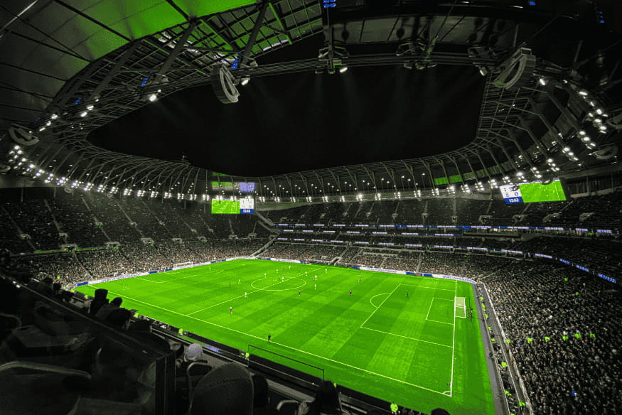 Estádio de futebol com cobertura moderna, iluminado durante jogo noturno com grande público.