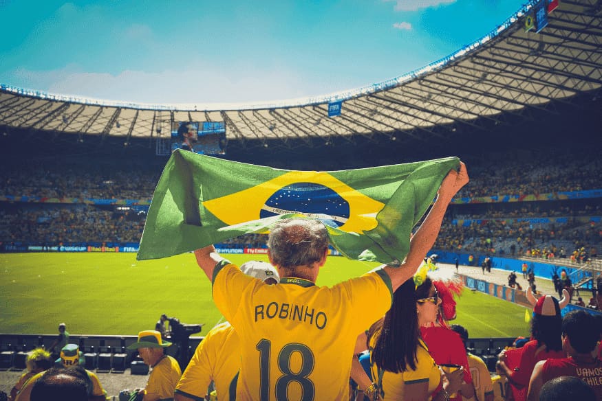 Homem com camisa da seleção e bandeira do Brasil aberta nas mãos durante jogo em estádio.