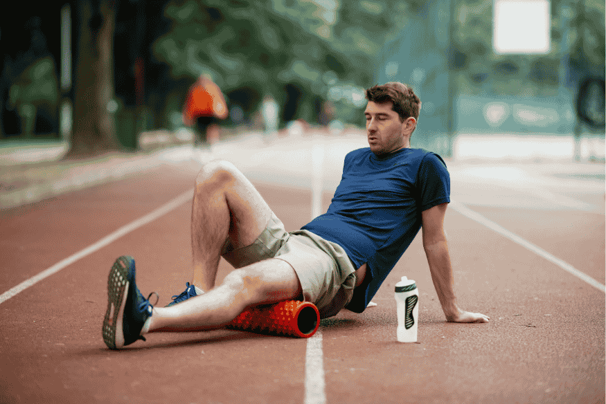 Corredor sentado em pista de atletismo usando foam roller para soltar a musculatura da coxa.