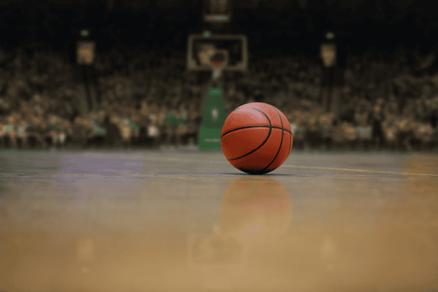 Bola de basquete em destaque no centro da quadra com torcida desfocada ao fundo.