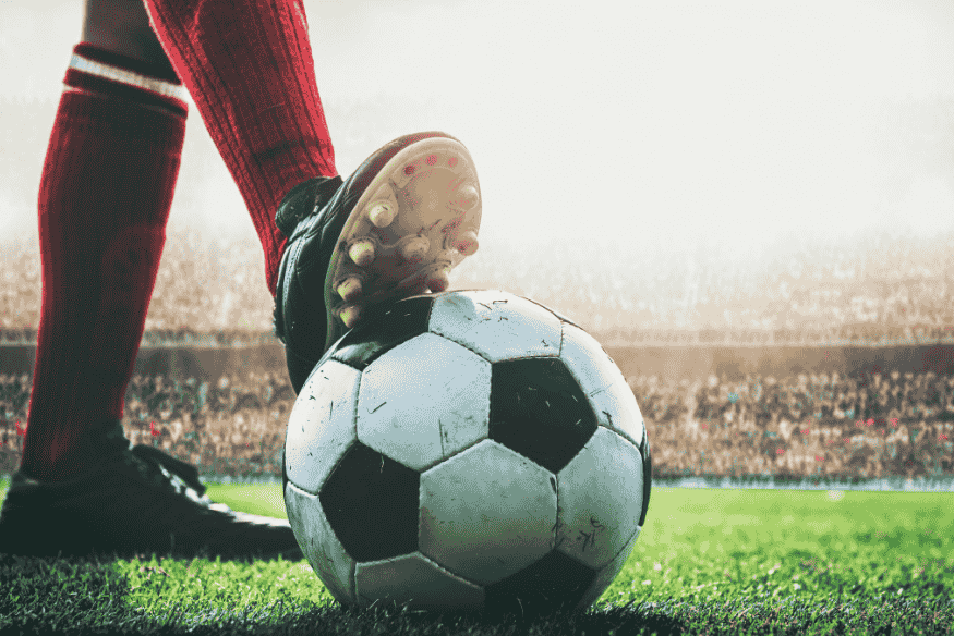 Close de jogador com chuteira vermelha pressionando uma bola de futebol em campo.