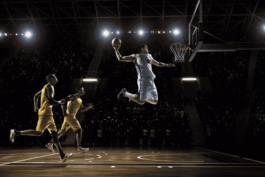 Jogador de basquete em uniforme branco voando em direção à cesta para uma enterrada durante jogo noturno.