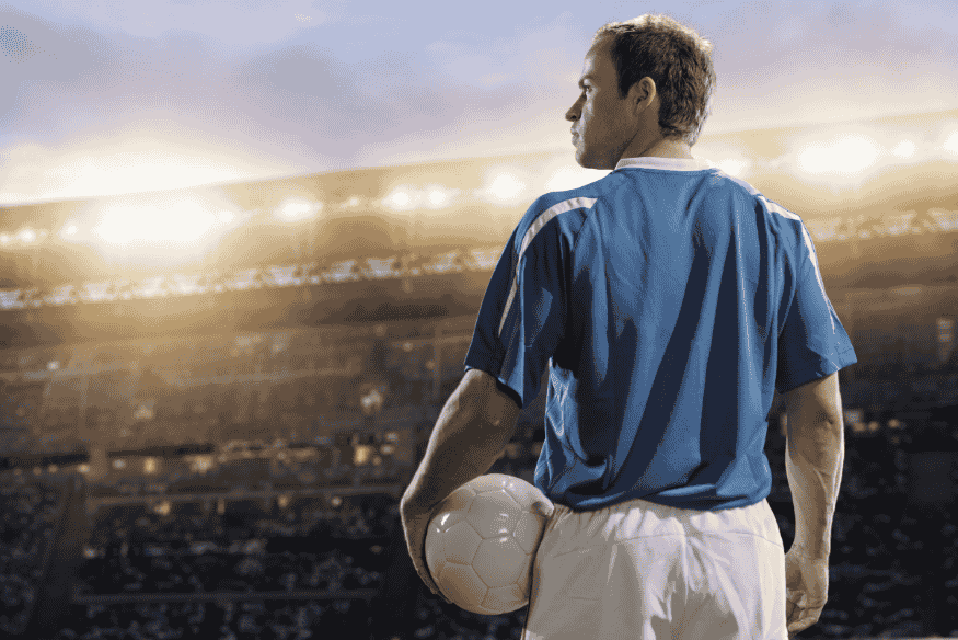 Jogador de futebol com bola nas mãos olhando para o campo sob luz de refletores.