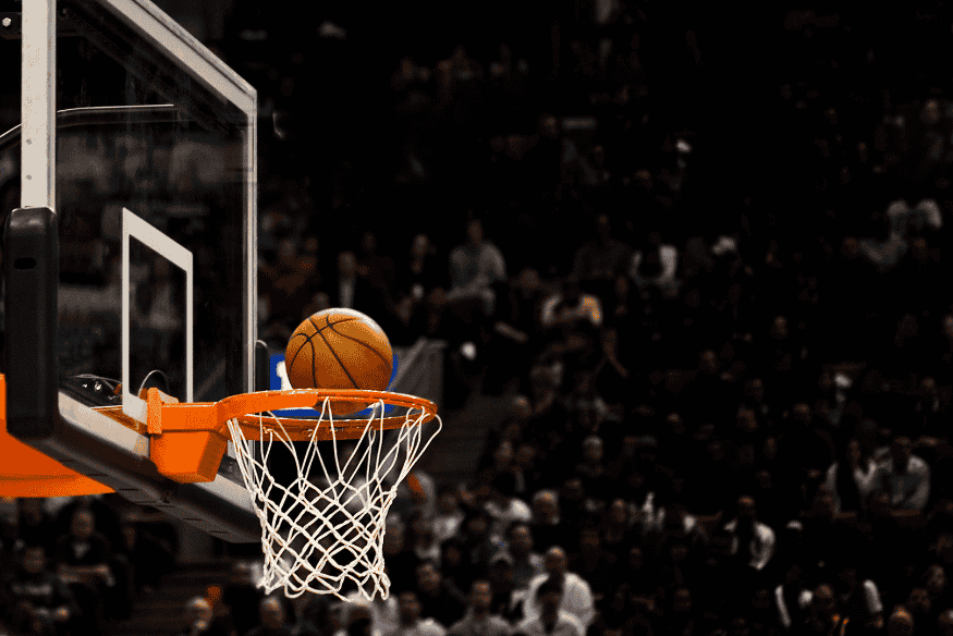 Bola de basquete prestes a cair na cesta durante jogo com torcida ao fundo.