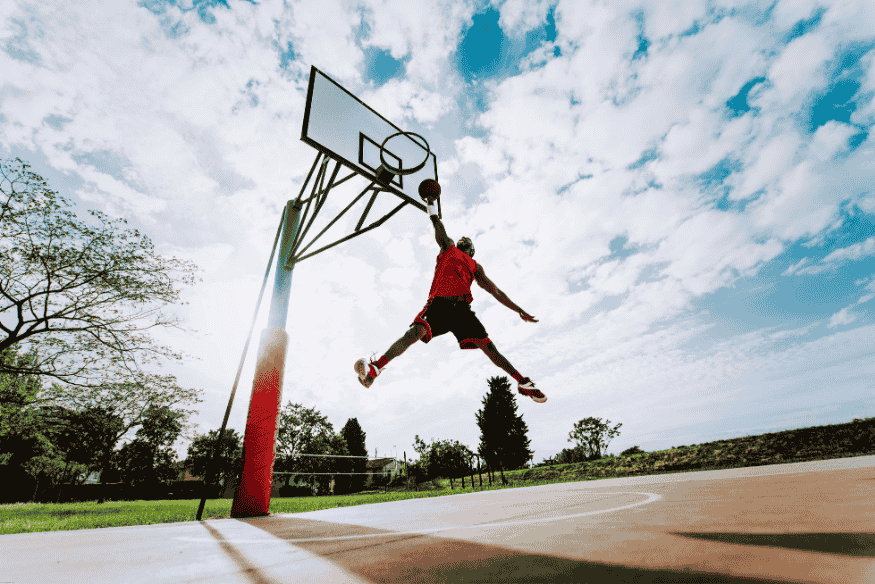 Atleta salta alto para uma enterrada em quadra de basquete ao ar livre com céu azul ao fundo.