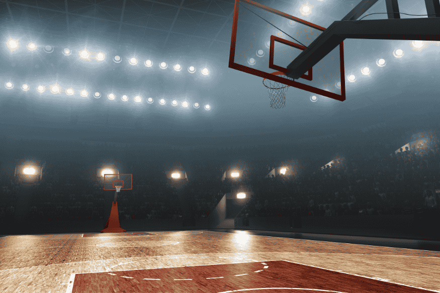 Quadra de basquete iluminada com tabelas e arquibancadas vazias antes do início da partida.