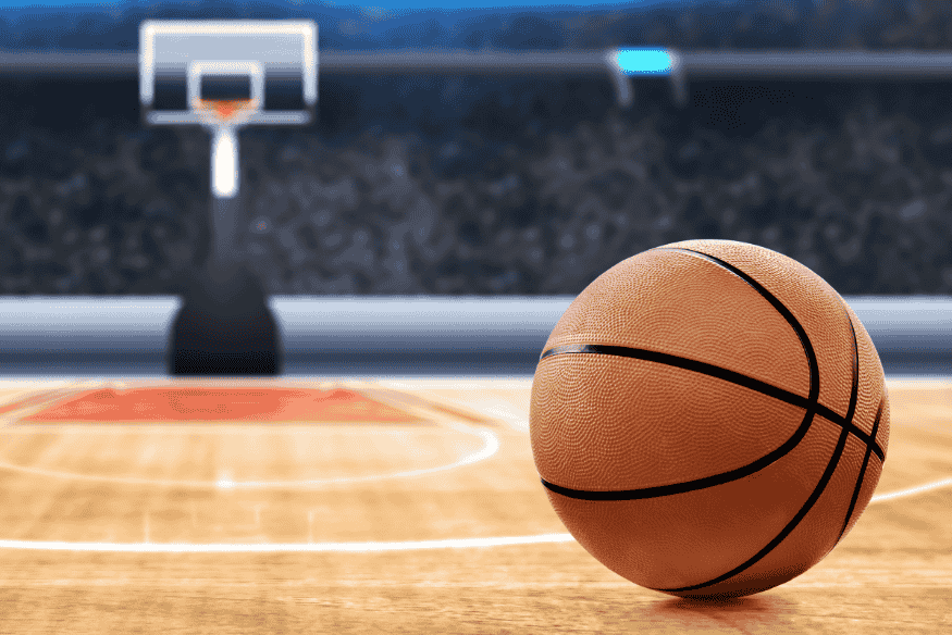 Bola de basquete em repouso sobre quadra indoor com tabela e cesta ao fundo.