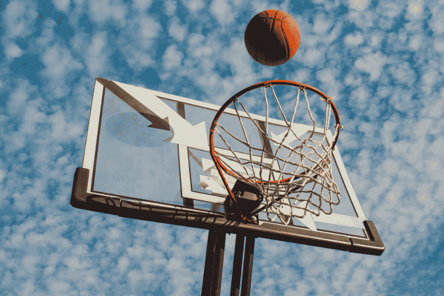 Bola de basquete no ar prestes a entrar na cesta em quadra externa com céu azul ao fundo.