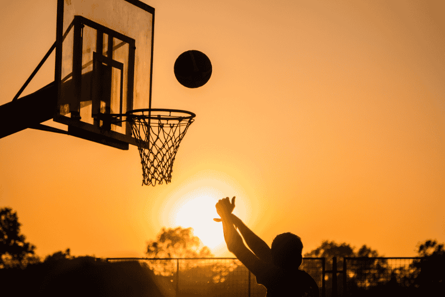 Pessoa arremessando bola de basquete em uma quadra externa ao pôr do sol.