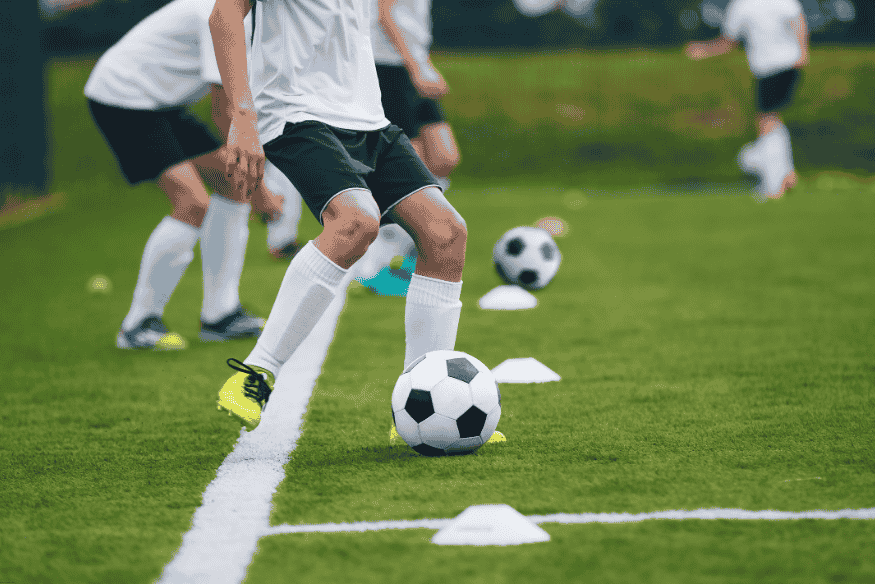 Jogadores de futebol fazendo treino de condução com cones e bola em campo demarcado.