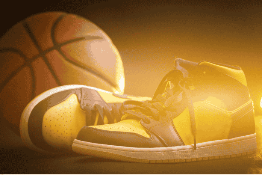 Par de tênis amarelos com detalhes pretos ao lado de uma bola de basquete em fundo iluminado.