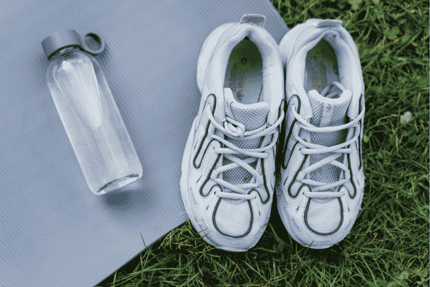 Par de tênis esportivos brancos ao lado de garrafa d’água sobre tapete de exercício no gramado.