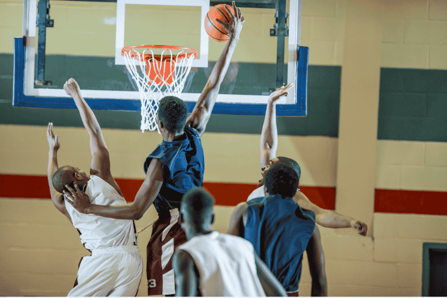 Jogador de basquete tentando uma enterrada enquanto é marcado por adversários sob a cesta.
