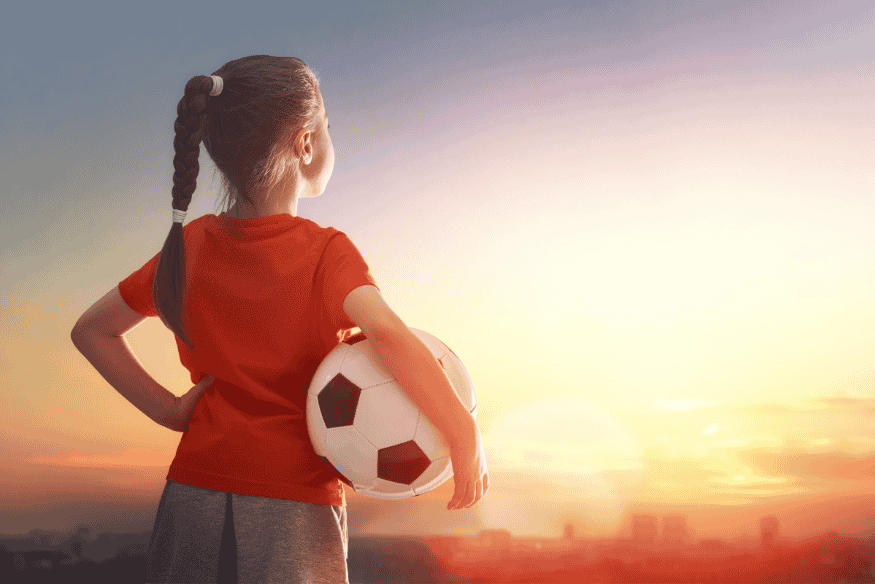 Menina segurando bola de futebol ao pôr do sol, olhando para o horizonte.