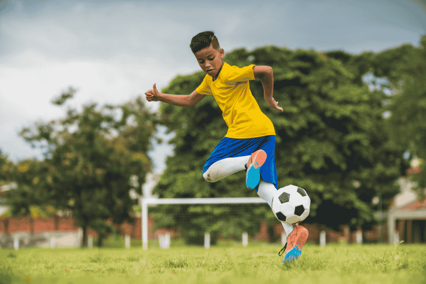 Criança com uniforme amarelo e azul treinando futebol em campo aberto.