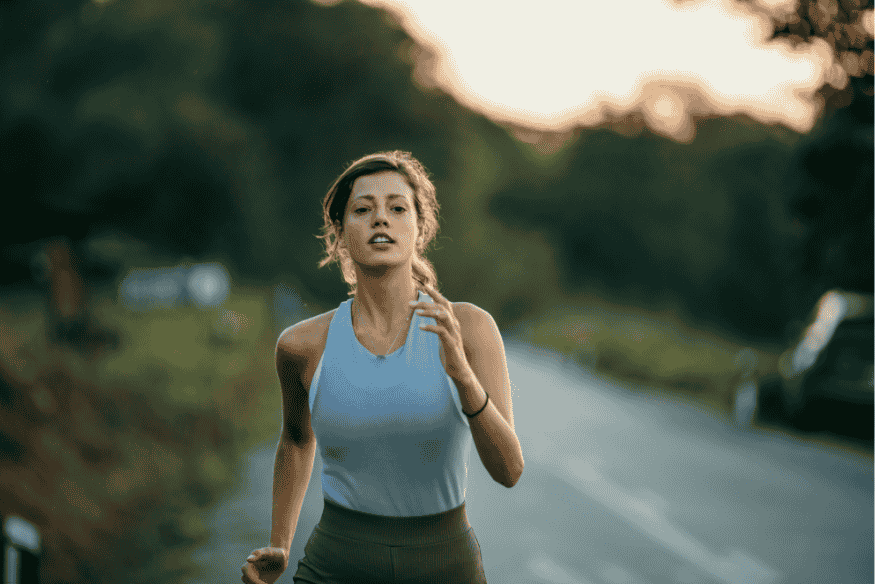Mulher correndo com intensidade em estrada arborizada, com expressão de foco e determinação.
