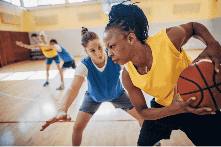 Mulher com colete amarelo protegendo a bola de basquete contra marcação intensa durante treino.