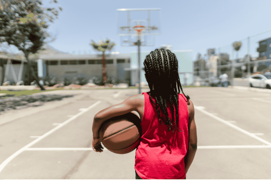 Garota com trancinhas segurando bola de basquete em quadra externa, olhando para a cesta.