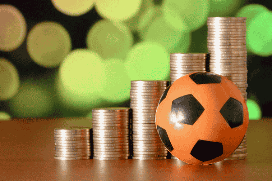 Bola de futebol laranja diante de pilhas crescentes de moedas, simbolizando finanças no futebol