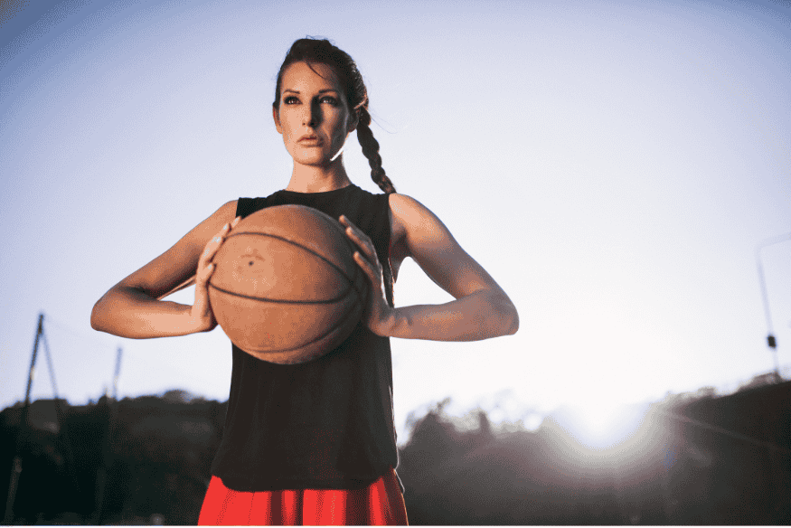 Jogadora de basquete segurando bola com expressão séria ao ar livre, ao pôr do sol.