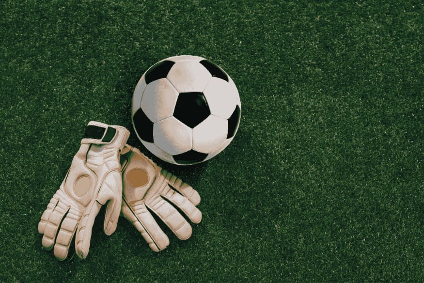 Luvas de goleiro ao lado de bola de futebol sobre gramado.