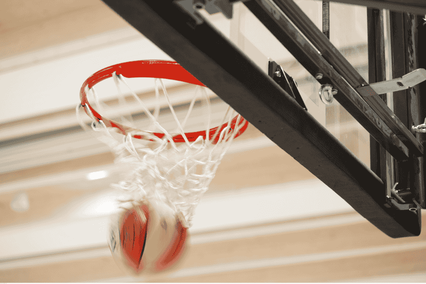 Bola de basquete passando pela rede do aro em movimento, com fundo de ginásio desfocado.