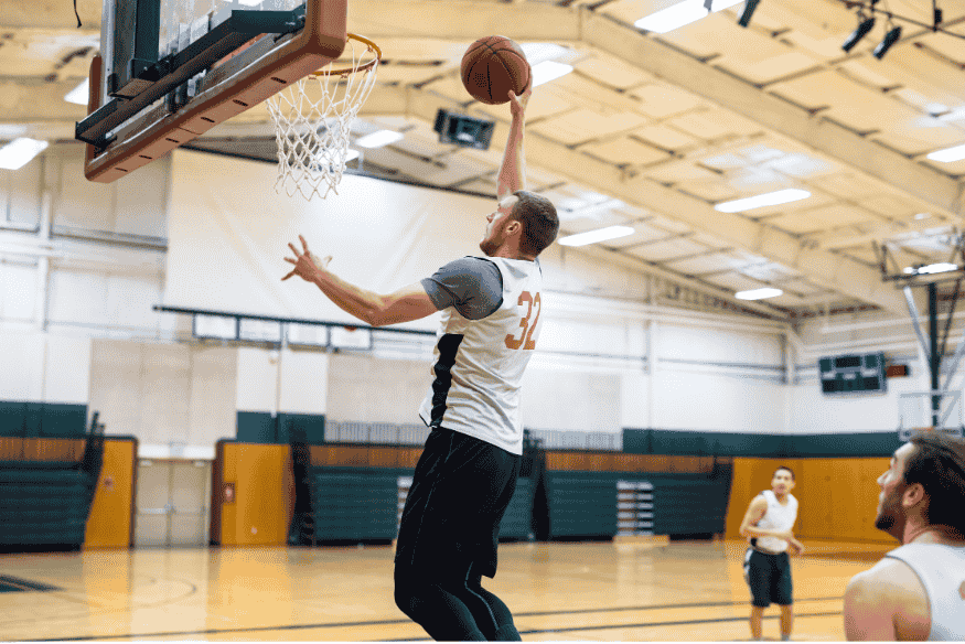 Jogador saltando para bandeja durante treino de basquete em quadra coberta com companheiros ao fundo.